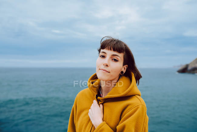 Frau in gelber warmer Jacke blickt in die Kamera, während sie vor dem plätschernden Meer und dem bewölkten Himmel in der Natur steht — Stockfoto