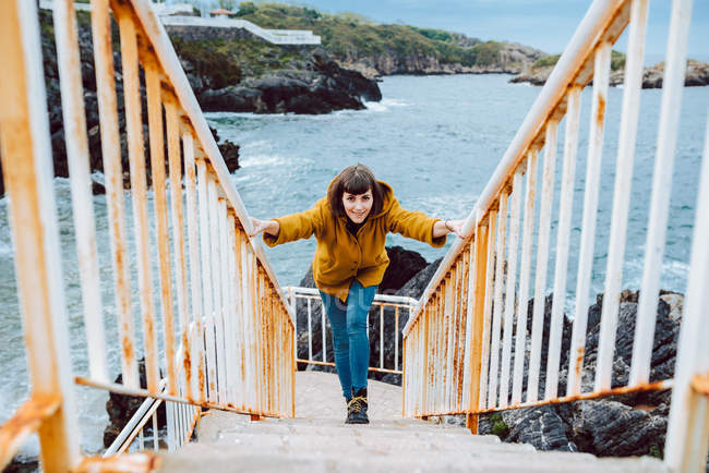 Молода жінка в жовтій куртці стоїть на іржавих сходах проти бухти з хвилястою морською водою — стокове фото