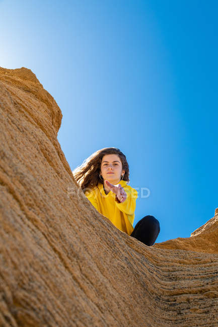 Porträt einer brünetten Frau in gelbem Sweatshirt, die Hand auf Sandsteinfelsen auf blauem Himmelshintergrund gibt — Stockfoto