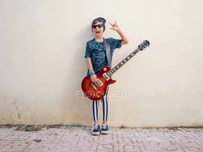 Cheeky active excited boy in colorful clothes playing guitar, showing two fingers up on background of white wall — Stock Photo