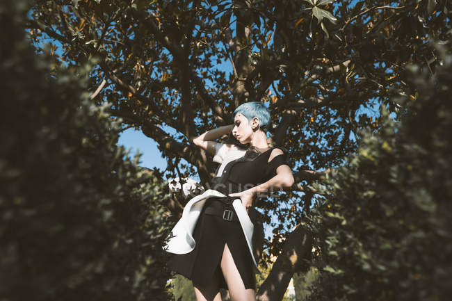 Von unten schaut junge Frau in futuristischem Kleid weg Kamera, während sie in Unschärfe neben Bäumen steht — Stockfoto