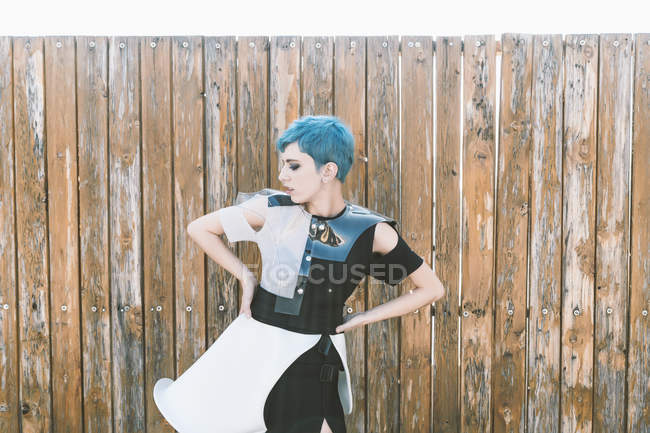Молодая женщина с короткими голубыми волосами в футуристическом платье и смотрит в сторону, стоя рядом с потрепанным деревянным забором — стоковое фото