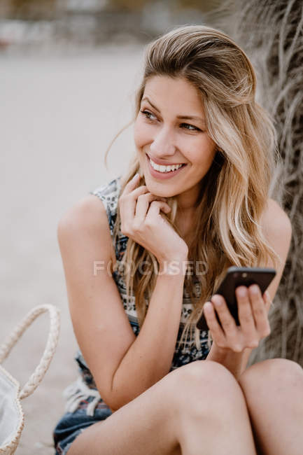 Joven atractiva hembra con el pelo largo relajándose en la orilla del mar y usando el teléfono móvil en el día de verano mientras mira hacia otro lado - foto de stock