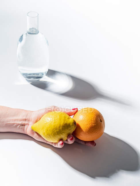Крупним планом жіноча рука тримає стиглий апельсин і лимон біля прозорої банки з водою на білому тлі — стокове фото