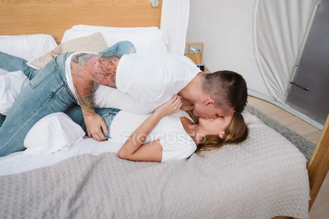 Side view of young romantic couple in white t-shirts and jeans kissing in bedroom of glamping — Stock Photo