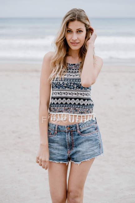 Mulher loira alegre em top colorido e calções jeans sorrindo e olhando para a câmera enquanto relaxa na praia — Fotografia de Stock