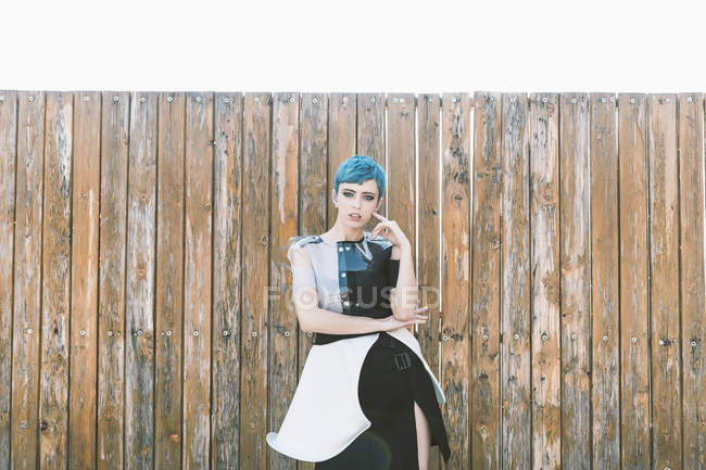 Молодая женщина с короткими голубыми волосами в футуристическом платье и смотрит в камеру, стоя рядом с потрепанным забором из древесины — стоковое фото