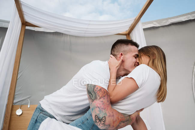 Vista laterale della giovane coppia romantica in t-shirt bianche e jeans inginocchiati e abbracciati in camera da letto di glamping — Foto stock