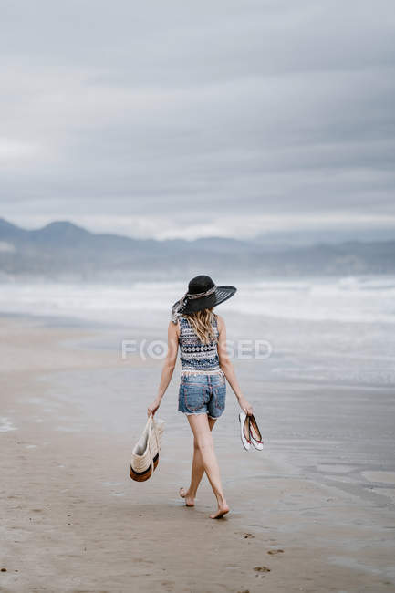 Rückansicht einer Frau mit schwarzem Hut, die eine Strandtasche in der Hand hält und den malerischen Blick auf das Meer genießt — Stockfoto