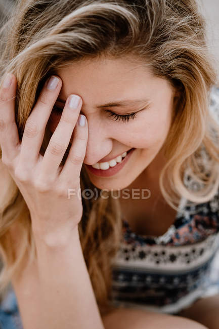 Retrato de cerca de una joven hermosa rubia con los ojos cerrados riéndose y cubriéndose la cara con la mano - foto de stock