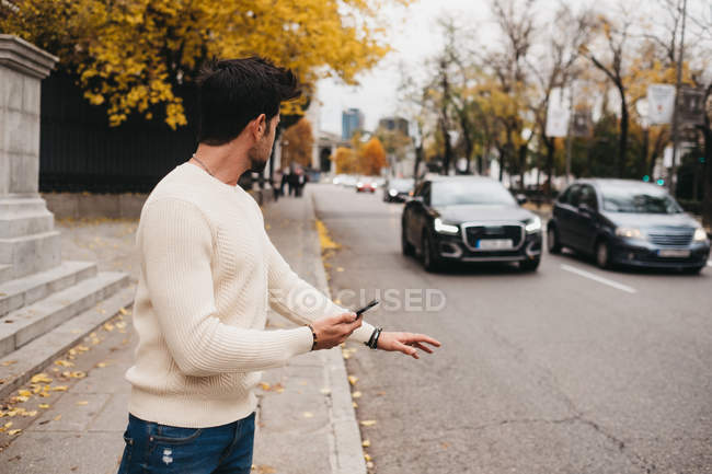 Giovane alla moda in piedi con il telefono cellulare sul ciglio della strada e la cattura di auto in autunno — Foto stock