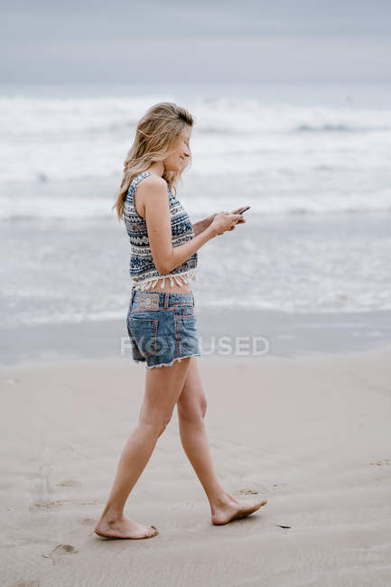Seitenansicht der jungen blonden fröhlichen Frau, die mit der Hand in der Tasche steht und mit dem Smartphone auf Meeresgrund spricht — Stockfoto