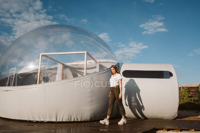 Jovem mulher bonita em t-shirt branca, calças e tênis posando por glamping transparente no fundo do céu azul — Fotografia de Stock