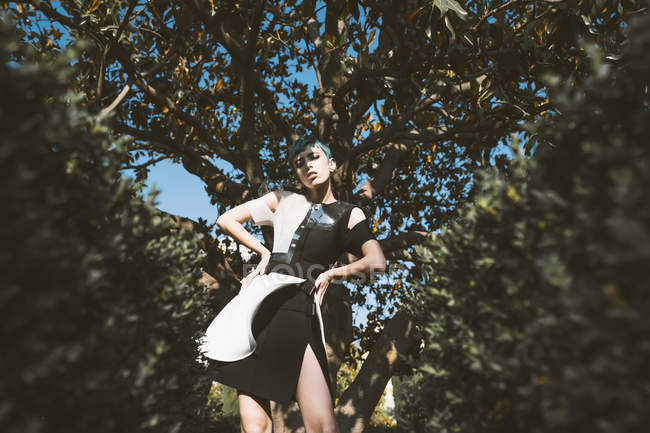 Von unten blickt junge Frau in futuristischem Kleid in die Kamera, während sie in Unschärfe neben Bäumen steht — Stockfoto