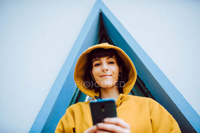 Frau justiert Kapuze aus gelbem warmen Mantel und schaut in Kamera, während sie in der Nähe des Gebäudes mit Dreiecksfenster Smartphone surft — Stockfoto