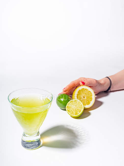 Female hand holding halved lemon and limes near glass of yellow lemonade drink on white background — Stock Photo