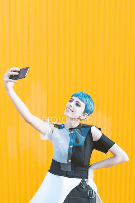 Молодая женщина в футуристическом платье смеется и делает селфи с телефоном, стоя напротив ярко-желтой стены — стоковое фото