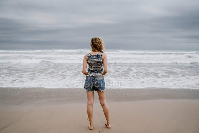 Back view of woman wearing top and shorts standing on sandy seashore — Stock Photo