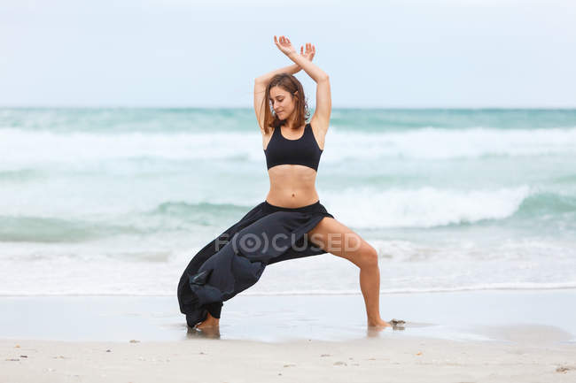 Молодая женщина в черном наряде танцует на песчаном пляже у моря — стоковое фото
