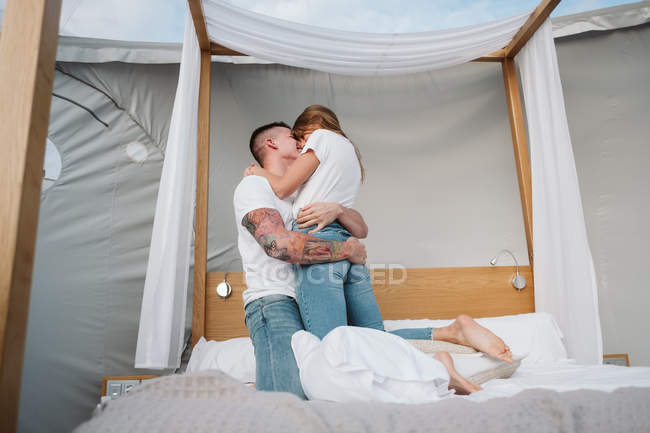 Вид сбоку на молодую романтическую пару в белых футболках и джинсах, стоящую на коленях и целующуюся в спальне глампинга — стоковое фото