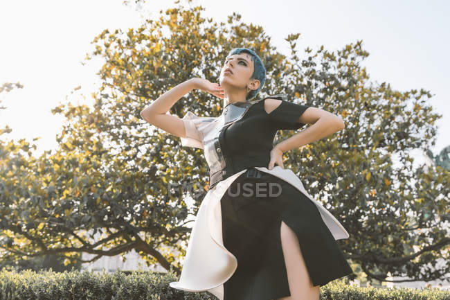 From below young woman in futuristic dress looking away while standing near trees in blur — Stock Photo