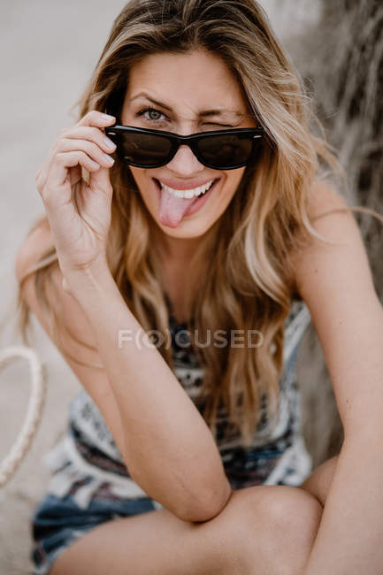 Gros plan portrait de femme blonde aux lunettes de soleil noires assise sur du sable et regardant la caméra sortir la langue et cligner des yeux — Photo de stock