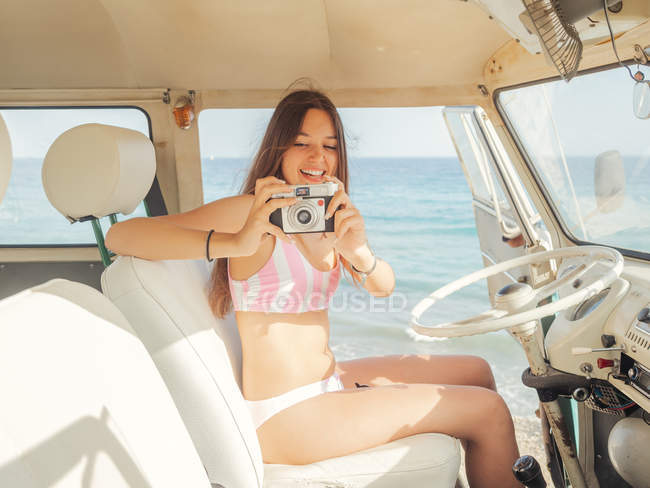 Mujer en traje de baño con la cámara sonriendo y tomando una foto en el asiento delantero blanco del coche en la playa en el día soleado - foto de stock