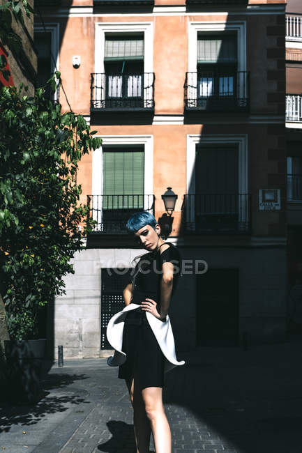 Молода жінка в футуристичній сукні стоїть руками на талії на вулиці проти старої будівлі на сонячному світлі — стокове фото