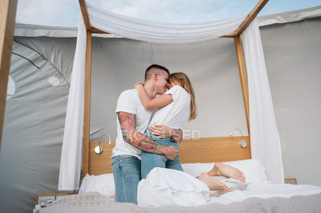 Вид сбоку на молодую романтическую пару в белых футболках и джинсах, стоящую на коленях и обнимающуюся в спальне глампинга — стоковое фото