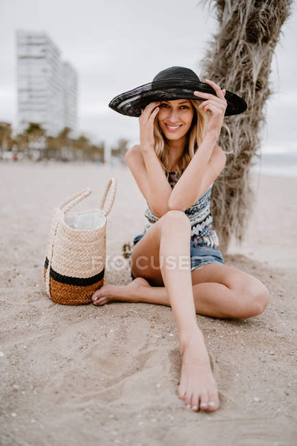 Mulher loira de chapéu preto sentado na areia com saco de verão e olhando para a câmera — Fotografia de Stock