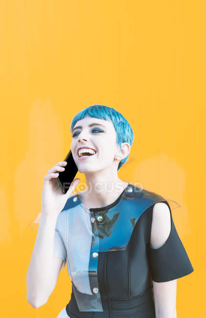 Jeune femme en robe futuriste riant et répondant à un appel téléphonique tout en se tenant contre un mur jaune vif — Photo de stock