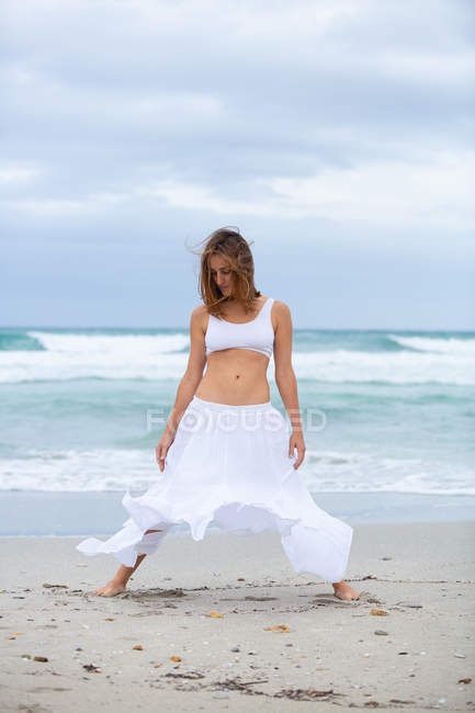 Attractive woman in white outfit dancing on sand near waving sea — Stock Photo