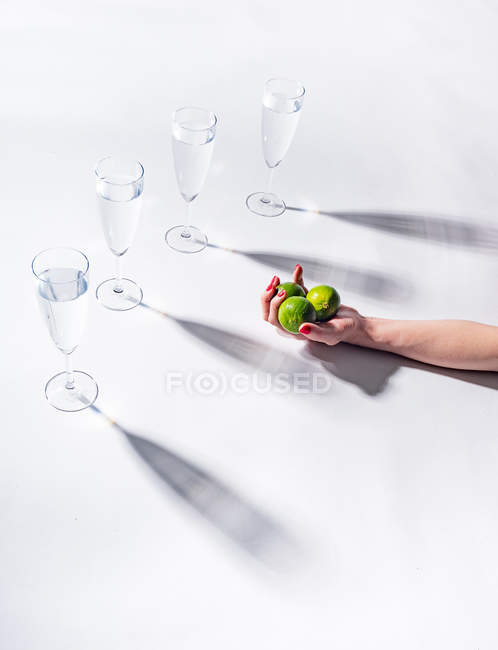 Женская рука держит зеленые спелые лаймы возле прозрачных стаканов воды на белом фоне — стоковое фото