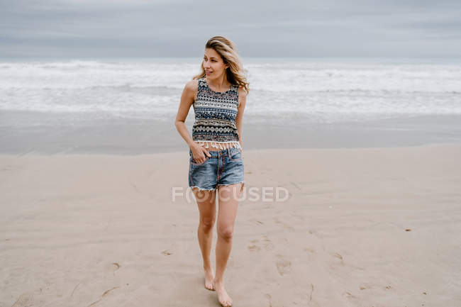 Mulher loira alegre em top colorido e calções jeans posando enquanto relaxa na praia — Fotografia de Stock