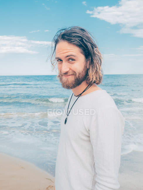 Side view of handsome happy adult bearded man standing relaxed by seashore looking at camera — Stock Photo