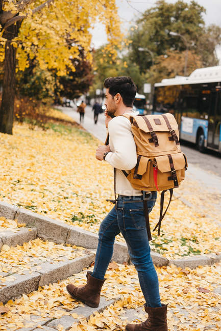 Красивый молодой человек с модной прической гуляет по осенним листьям и говорит по смартфону днем — стоковое фото