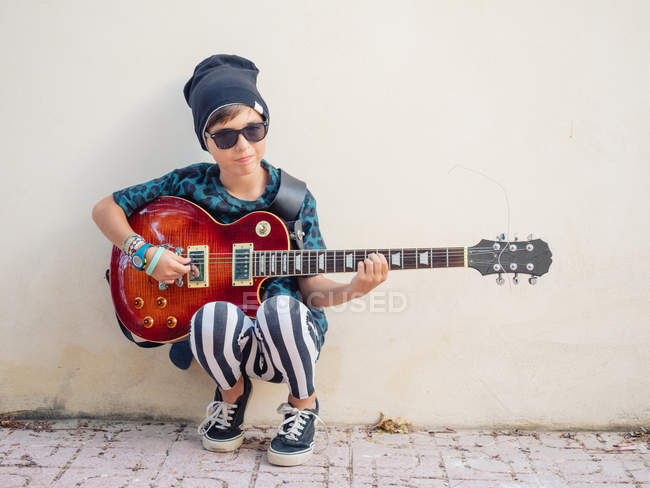 Нахабний активний збуджений хлопчик в барвистому одязі грає на гітарі, спираючись на тлі білої стіни — стокове фото