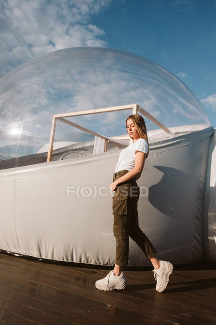 Jeune belle femme en t-shirt blanc, pantalon et baskets posant par glamping transparent sur fond bleu ciel — Photo de stock