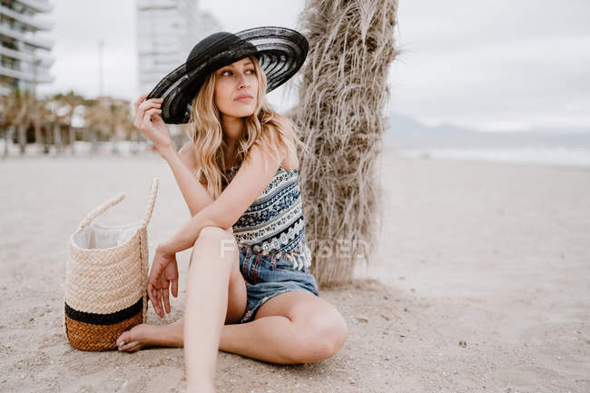 Femme blonde en chapeau noir assis sur le sable avec sac d'été et regardant loin — Photo de stock