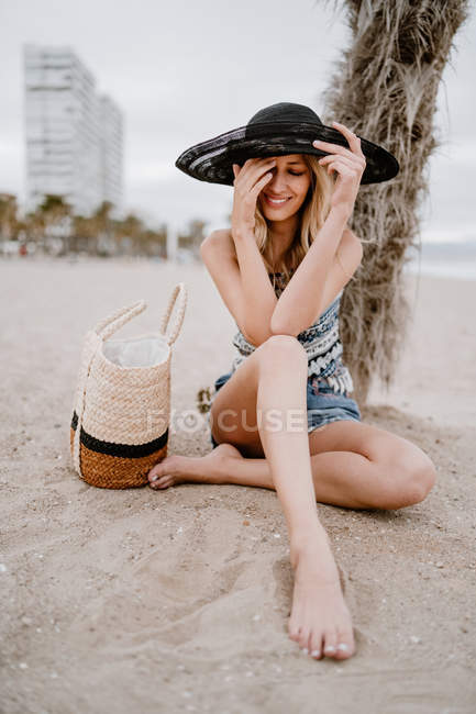 Donna bionda in cappello nero seduta sulla sabbia con borsa estiva ad occhi chiusi — Foto stock