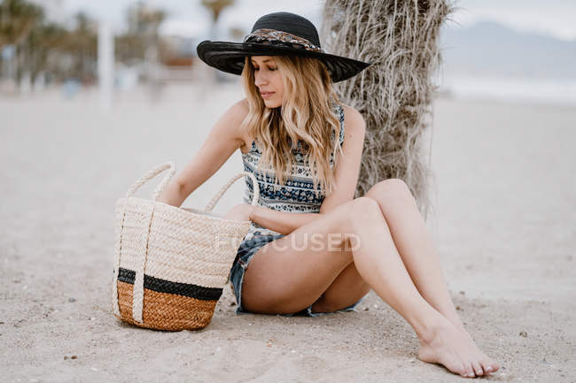 Блондинка в чорному капелюсі сидить на піску з літньою сумкою і дивиться вниз — стокове фото