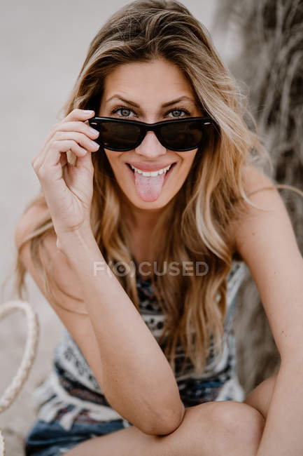 Крупный план портрета блондинки в черных солнцезащитных очках, сидящей на песке, смотрящей в камеру и торчащей языком — стоковое фото