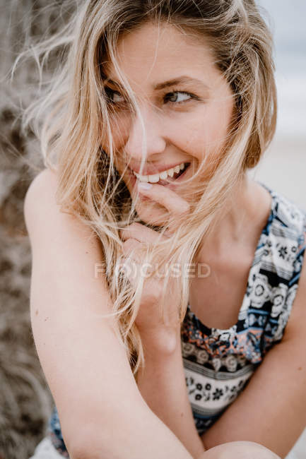Mujer rubia en la parte superior mordiendo dedo índice y sonriendo a la cámara en el fondo de la naturaleza - foto de stock