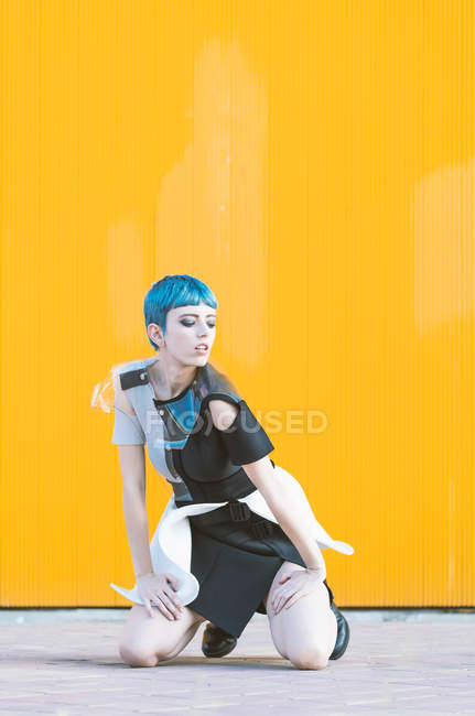 Молода жінка в модному футуристичному вбранні, сидячи на колінах на тротуарі до яскраво-жовтого муру. — стокове фото