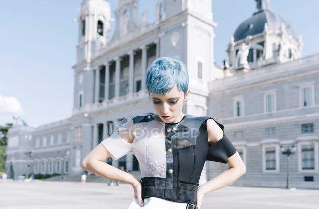 Mujer joven en futurista de moda con las manos en la cintura y el tacto mientras está de pie en la calle de la ciudad en el día soleado - foto de stock