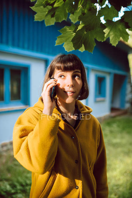 Young woman in casual outfit smiling and talking by smartphone while standing on tiled path outside lovely cottage on autumn day in countryside — Stock Photo