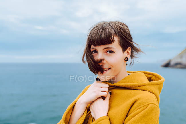 Mulher de casaco quente amarelo olhando para a câmera enquanto está contra o mar ondulante e céu nublado na natureza em um dia ventoso — Fotografia de Stock