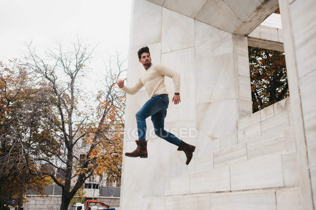 Jovem na moda em jeans e camisola branca correndo no ar no edifício branco — Fotografia de Stock