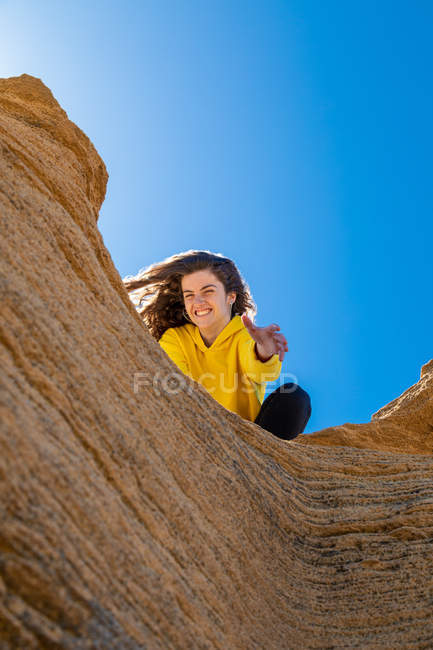 Портрет брюнетки в жёлтой толстовке, протягивающей руку на скале из песчаника на голубом фоне неба — стоковое фото