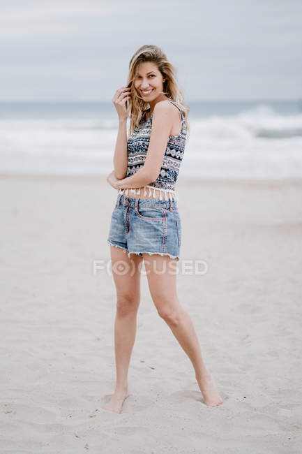 Mulher loira alegre em top colorido e calções jeans sorrindo e olhando para a câmera enquanto relaxa na praia — Fotografia de Stock